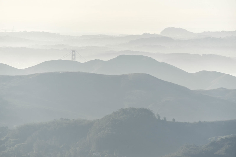 Rows of hills shrouded by fog; the top of the Golden Gate Bridge is visible in the background